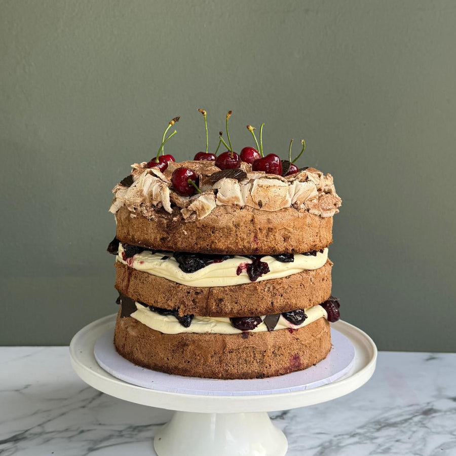 A tall three-layer Black Forest sponge cake filled with whipped cream, cherry compote, topped with rustic baked meringue crown and fresh cherries, sitting on a white cake stand 