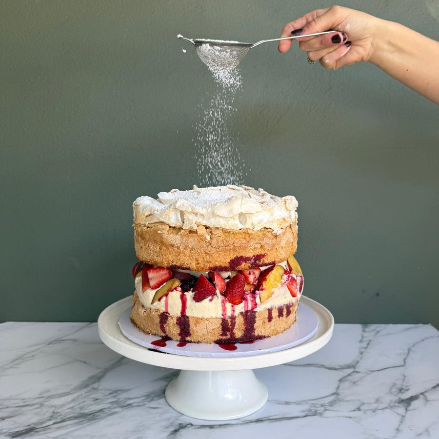 Hand dusting icing sugar over a layered sponge cake filled with summer berries, cream, market peaches and berry syrup om a white cake stand