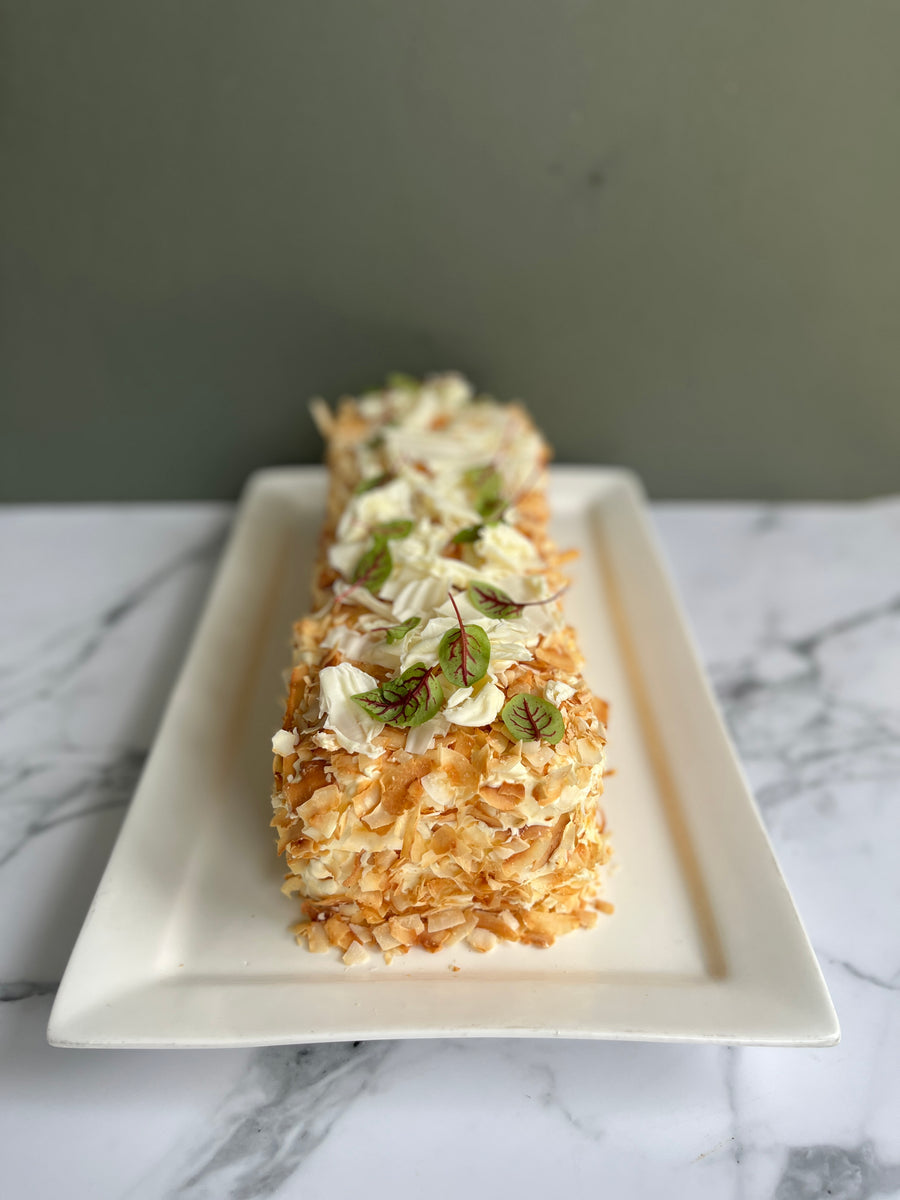 Rectangular pavlova displayed on a rectangular platter. The cake is covered with toasted coconut and topped with tempered white chocolate, on a marble surface with a green background. Premium hadcrafted French dessert for christmas from Millstone Patisserie 